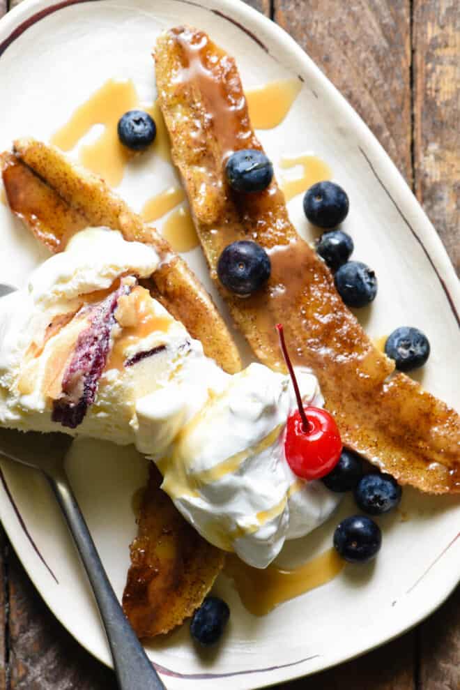 Small oblong shaped white plate topped with a frozen dessert made with bananas, ice cream, blueberries, caramel sauce and a cherry.
