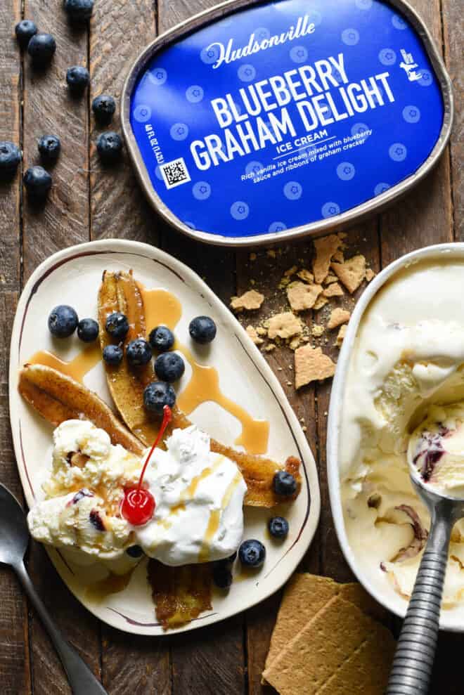 Small oblong shaped white plate topped with browned bananas, ice cream, blueberries, caramel sauce and a cherry. A container of Blueberry Graham Delight ice cream is next to plate.