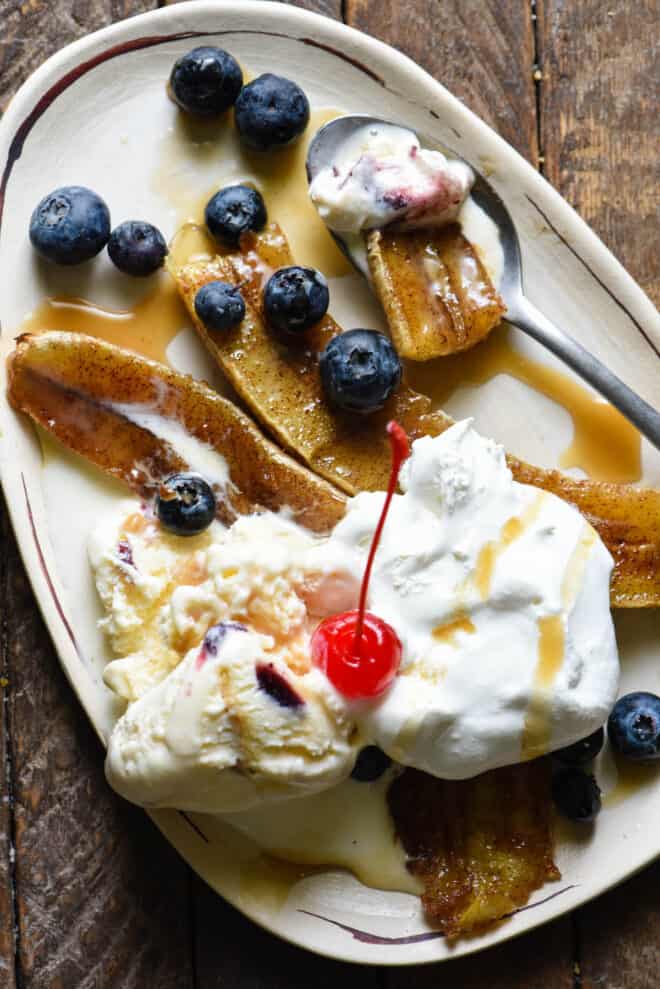 Small oblong shaped white plate topped with a broiled banana, ice cream, blueberries, caramel sauce and a cherry.