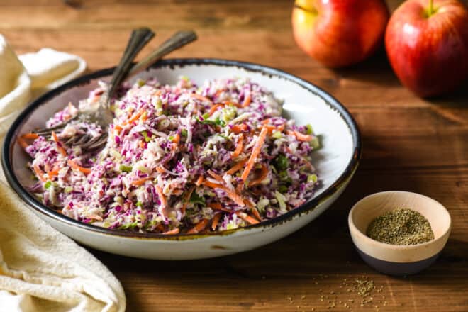 Shallow white bowl filled with coleslaw with celery seed, with smaller bowl of celery seed next to it, and apples in background.