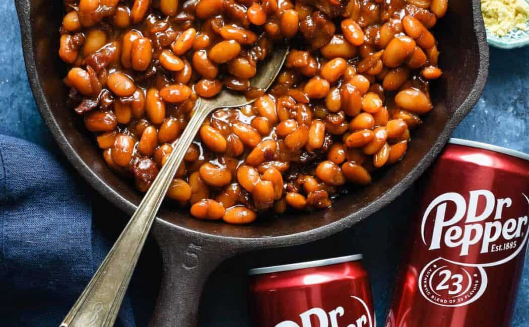 Cast iron skillet filled with baked beans with cans of Dr. Pepper next to skillet.