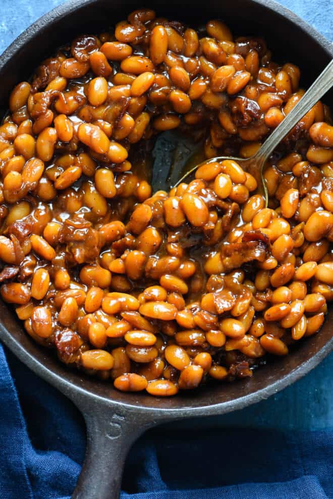 Cast iron skillet filled with Dr. Pepper baked beans, with spoon dishing out some beans.