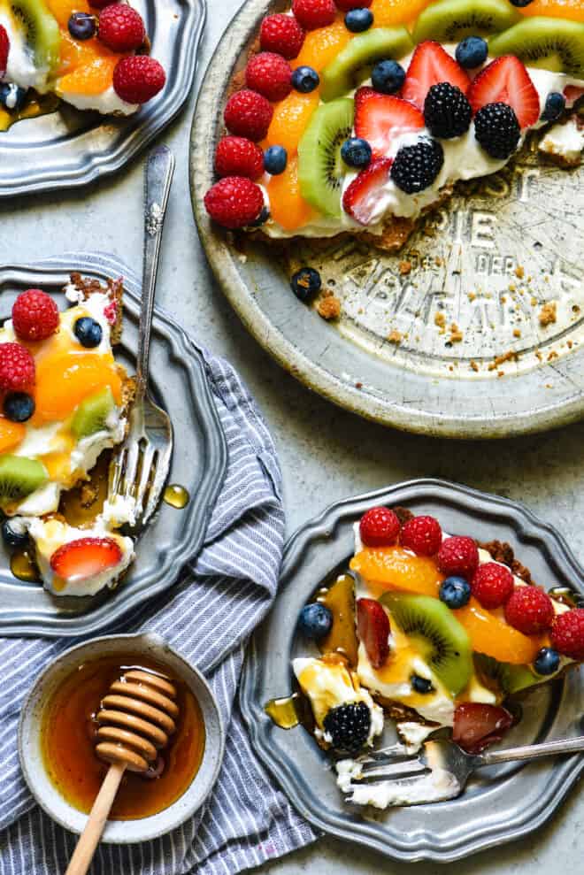 Antique pie pan with half of a creamy pie topped with fruit, alongside plates with pie slices topped with honey.