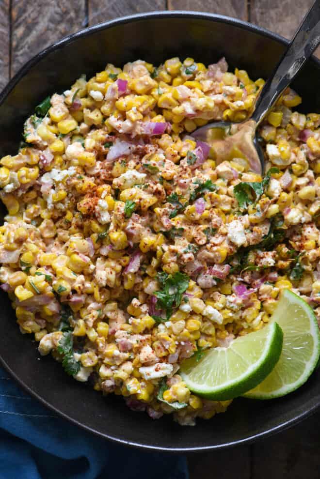 Corn dish with cheese and red onions in a black bowl, garnished with lime wedges.