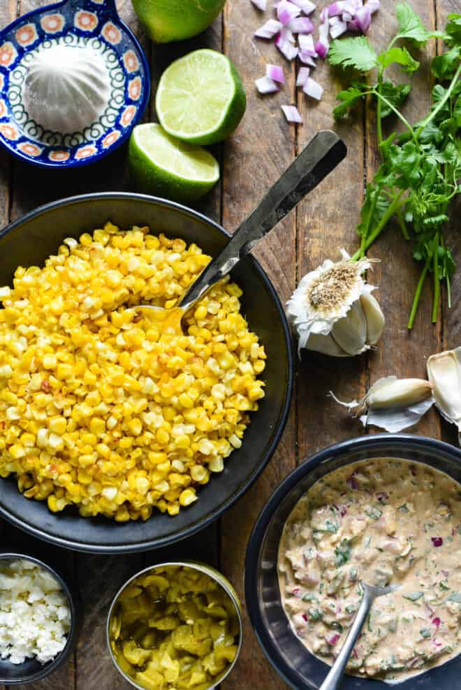 Ingredients for elote salad on a wooden surface, including corn kernels, creamy sauce, feta cheese, chopped red onion and garlic.