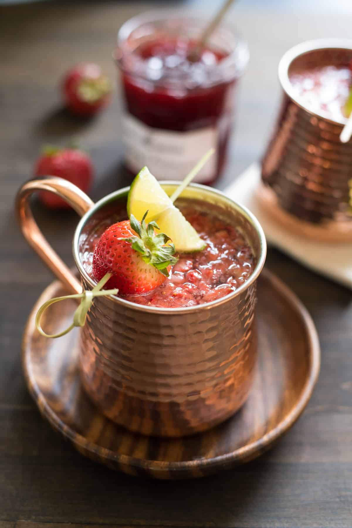Hammered copper mug filled with strawberry Moscow mule, garnished with a skewer with a strawberry and lime wedge. Mug is resting on small wooden plate.