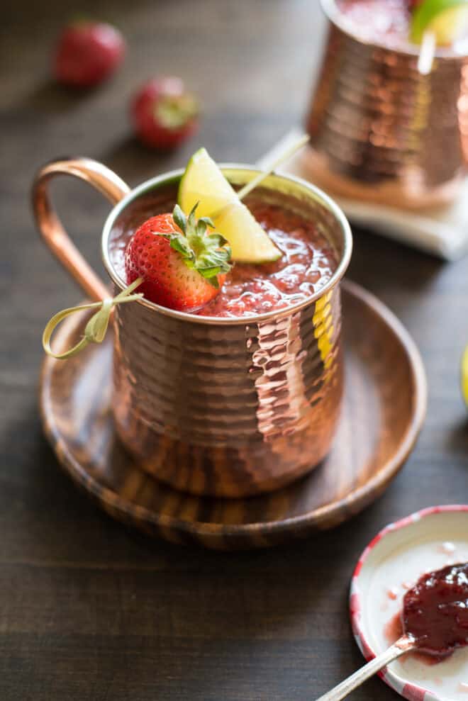 Hammered copper mug filled with strawberry Moscow mule, garnished with a skewer with a strawberry and lime wedge. Mug is resting on small wooden plate.