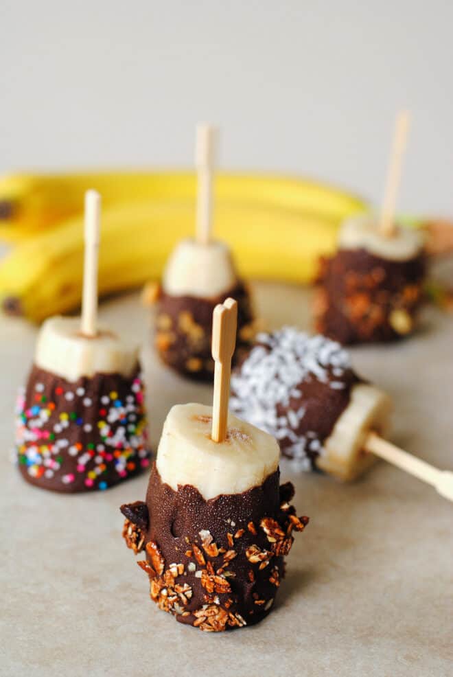 Toothpicks skewered with frozen chocolate bananas sprinkled with toppings like granola, sprinkles and coconut.