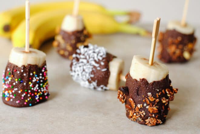 Small chocolate covered bananas sprinkled with toppings, on toothpicks.