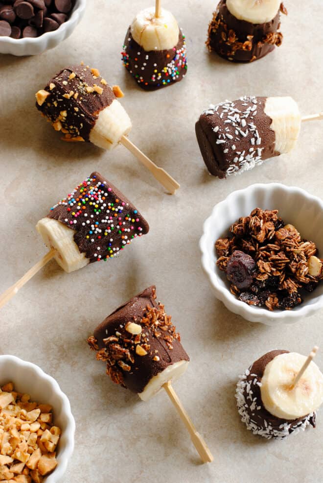 Small frozen chocolate covered bananas on a beige surface, with small bowls of toppings like granola and nuts.