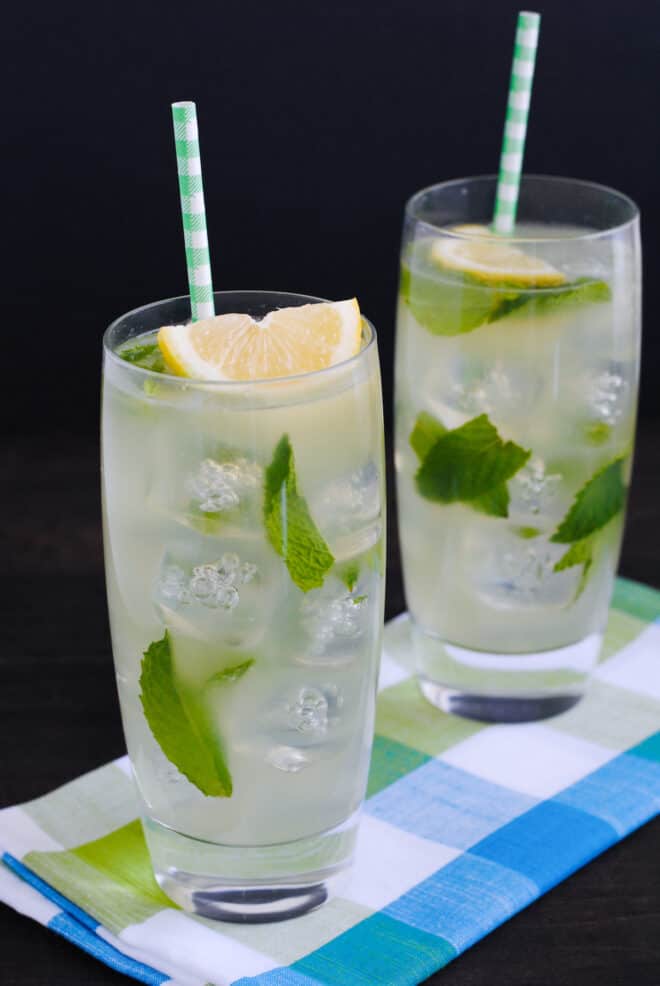 Two tall glasses of lemon drink garnished with lemon wedges, mint leaves and paper straws.