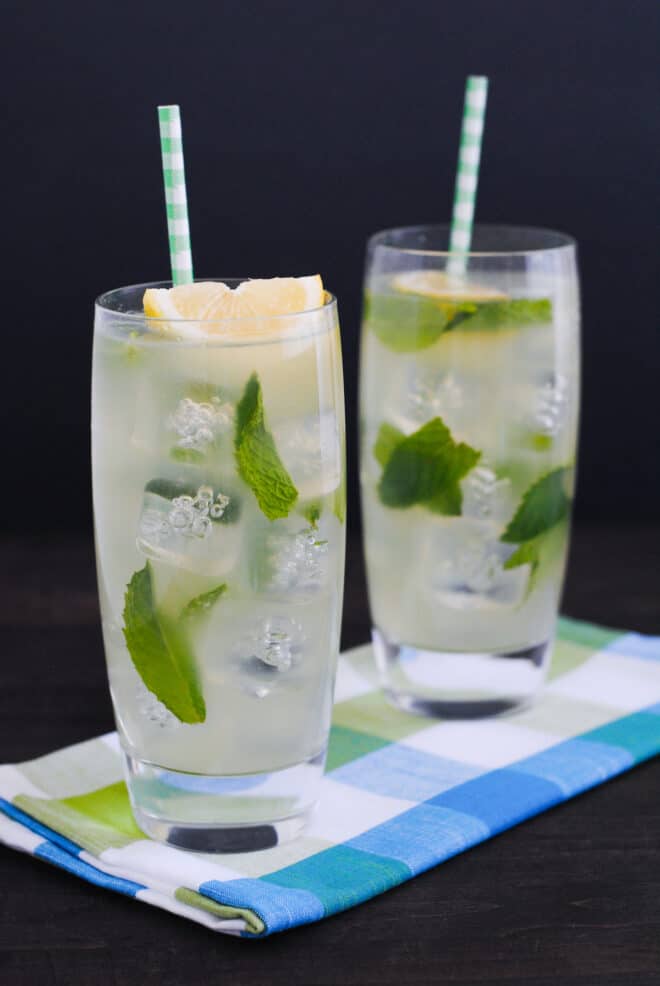 Two tall glasses filled with ice, honey lemonade, mint leaves, lemon wedges and paper straws.