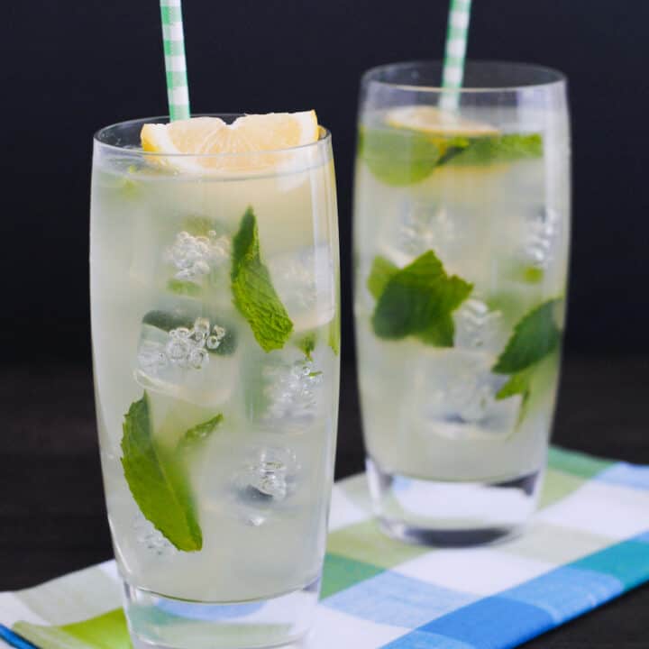 Two tall glasses filled with ice, honey lemonade, mint leaves, lemon wedges and paper straws.