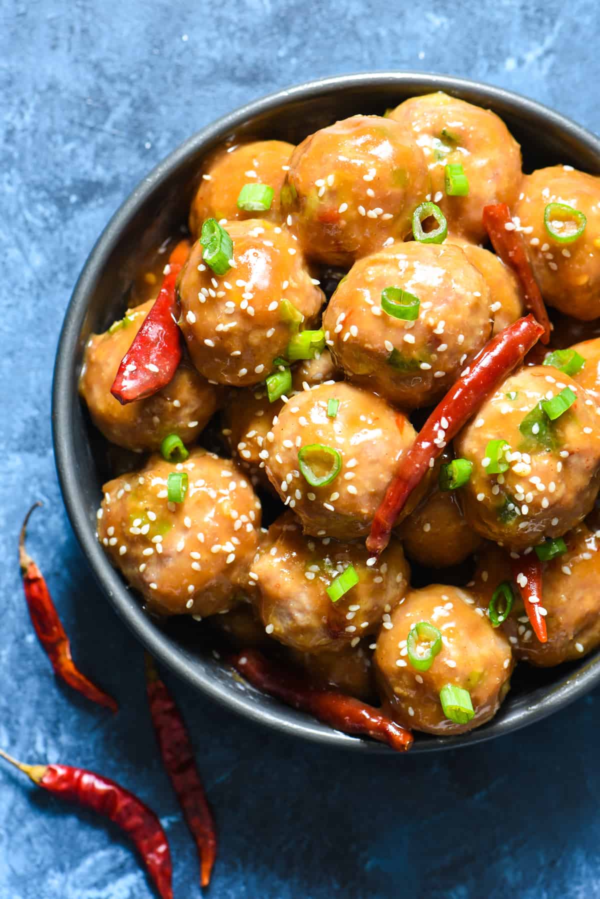Round bowl of General Tso's meatballs topped with green onions, sesame seeds and dried chiles on a blue background.