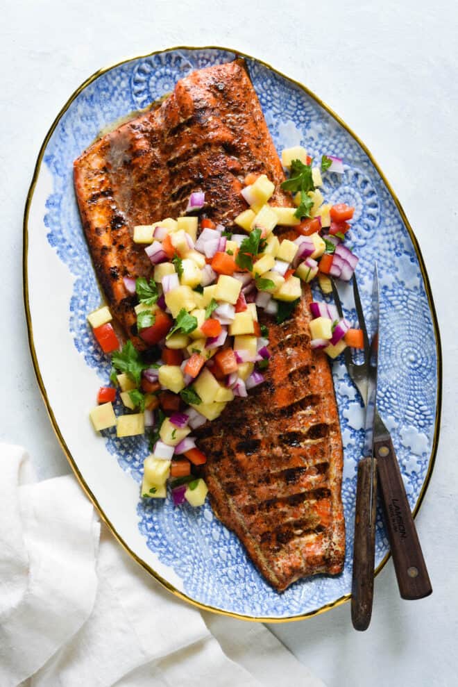 Decorative blue and white platter topped with a large piece of grilled fish with skin and chopped pineapple salsa.