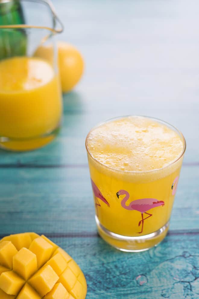 Glass with flamingo printed on it on teal table, filled with an orange hued beverage.