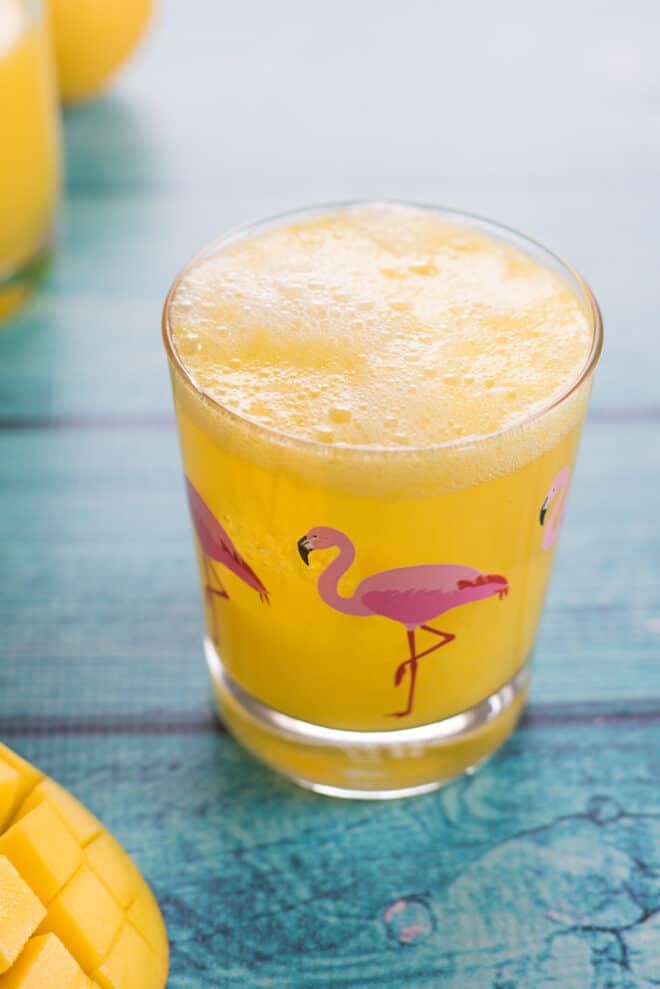 Glass with flamingo printed on it on teal table, filled with an orange hued beverage. 