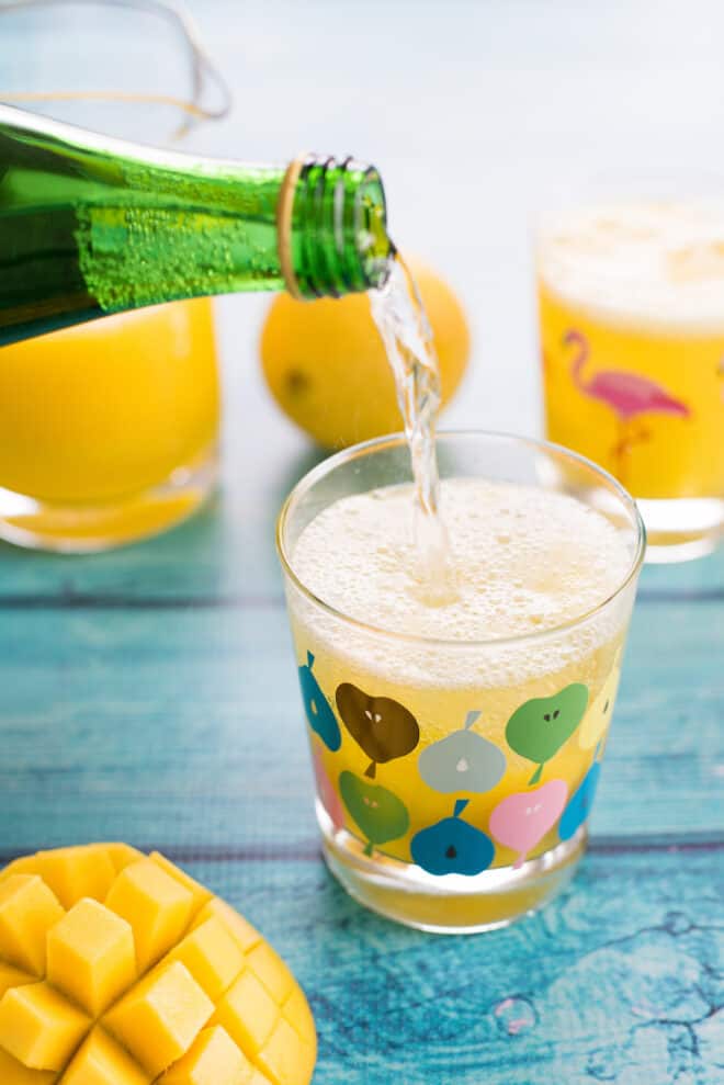 Glass of sparkling mango lemonade on teal table, with fruit and bottles of juice and water in background and foreground, with bottle pouring fizzy water into glass.