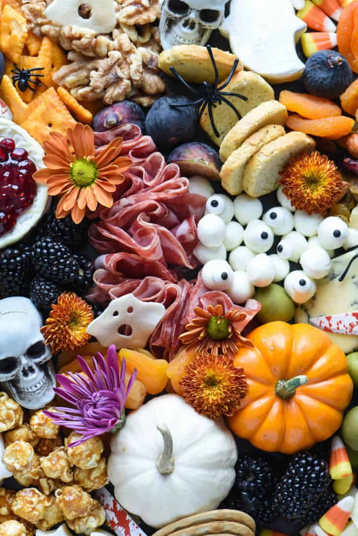 A spooky charcuterie board made with salami, mini mozzarella cheese, blackberries and dried apricots. Pumpkins and fall flowers decorate the tray.