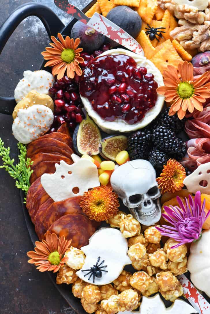 Halloween cheese board made with French cheese round topped with red jam, pumpkin cookies, pepperoni, crackers, walnuts, figs, and spooky decorations.