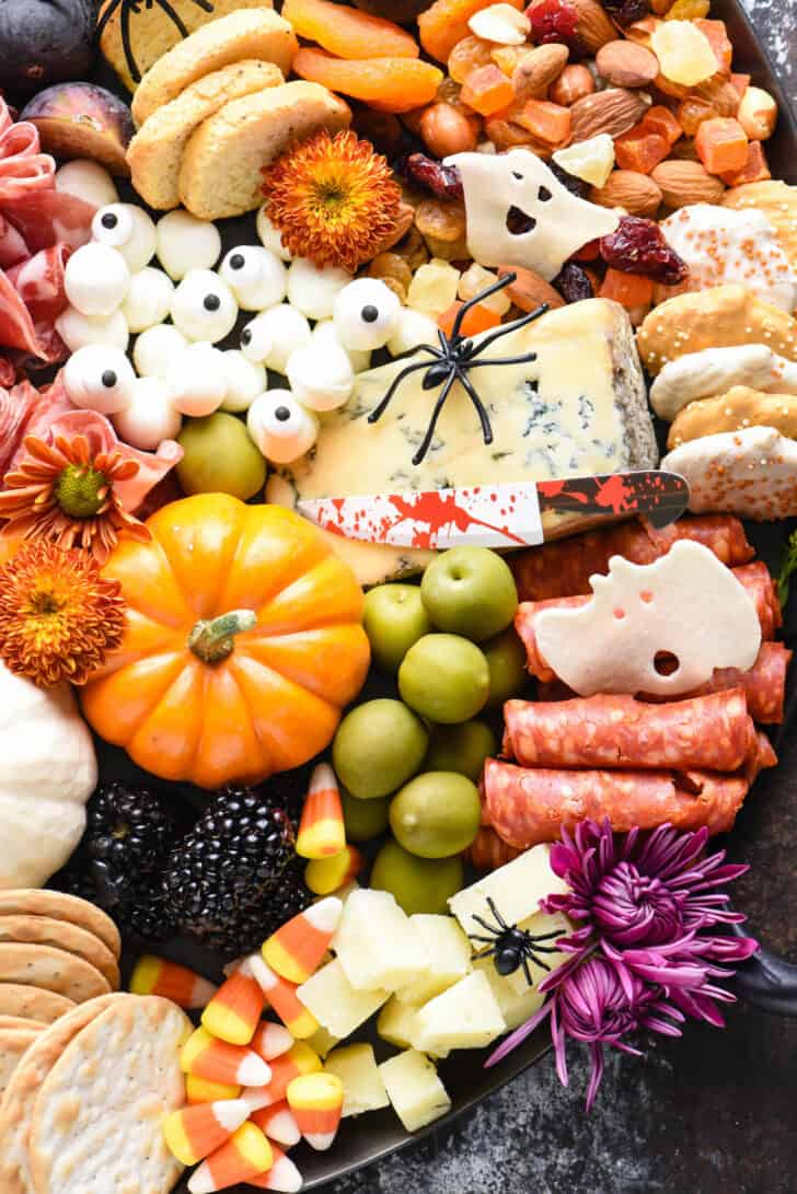 A spooky meat and cheese tray made with blue cheese, mini mozzarella balls, crackers, olives, mini pumpkins, cookies and trail mix. Plastic spiders on cheese.