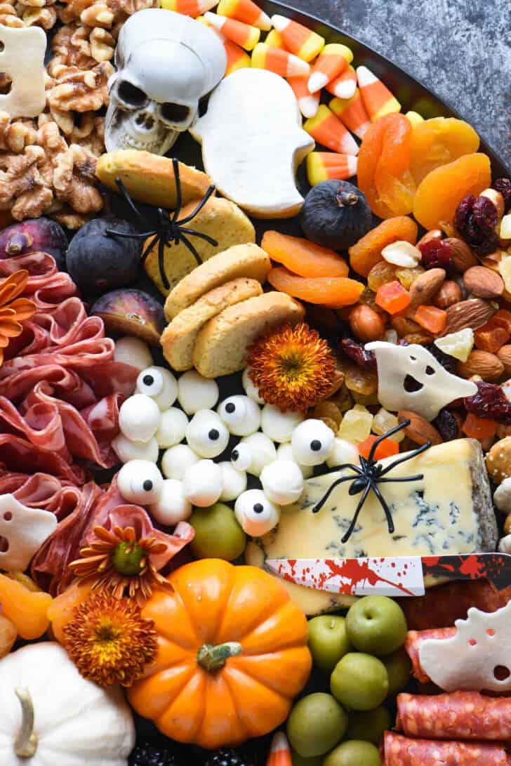 Fall themed snack platter made with blue cheese, mini mozzarella balls, cured meat, figs, crackers, olives, mini pumpkins, cookies and trail mix. Plastic spiders and skulls decorate the tray.