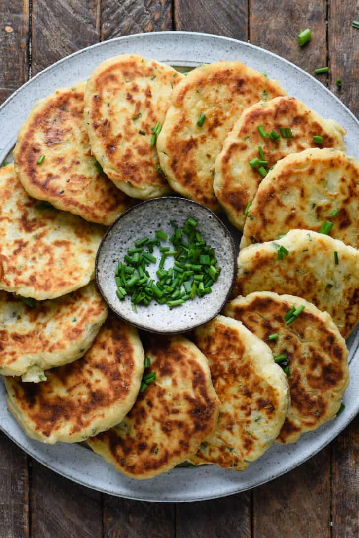 Gray plate topped with mashed potato cakes arranged in a circle, with a small bowl of chives in center of circle.