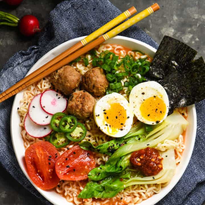 Ramen noodles with tomato broth in a white bowl, topped with meatballs, a soft boiled egg, and a variety of vegetable garnishes.