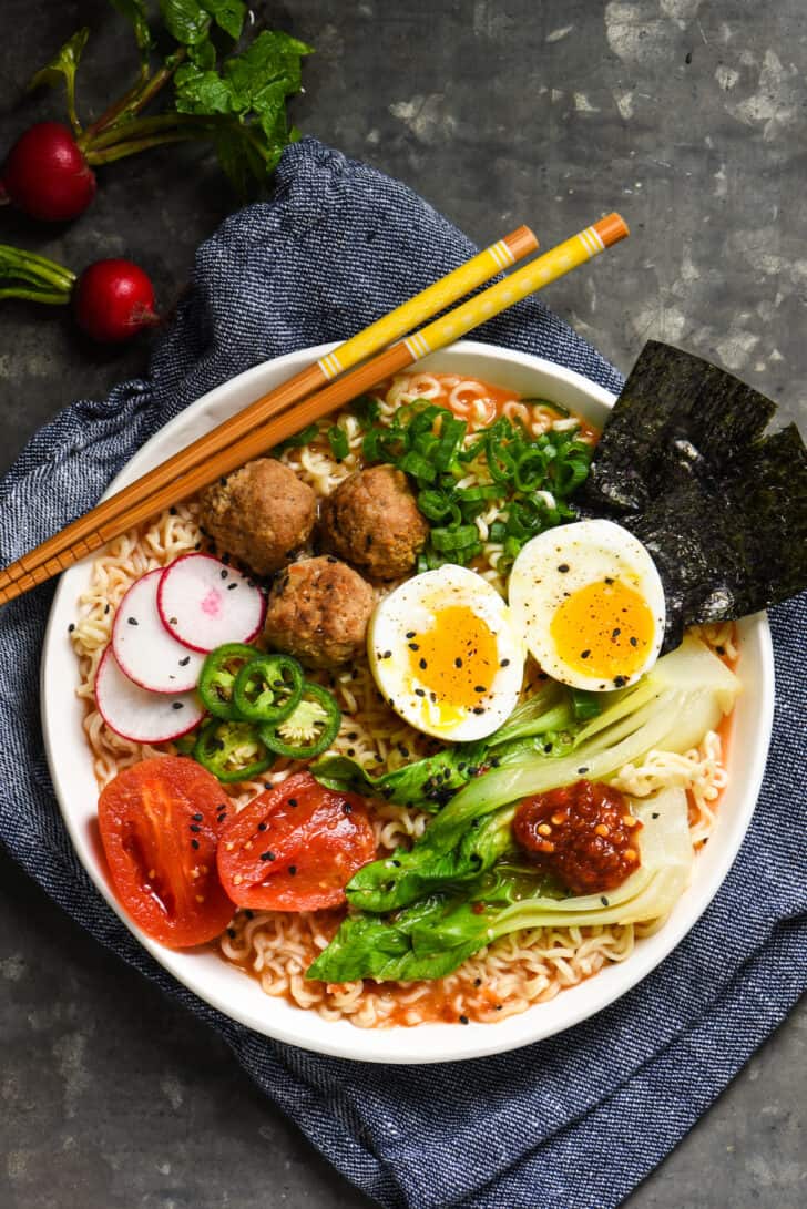 Ramen noodles with tomato broth in a white bowl, topped with meatballs, a soft boiled egg, and a variety of vegetable garnishes.