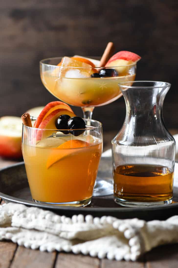 A tray of apple cider bourbon cocktails in various glasses, with a carafe of whiskey as well.