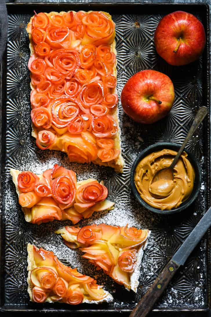 Apple tart made with decorative roses made from apples, cut into pieces on vintage baking pan, with bowl of peanut butter and fresh apples nearby.