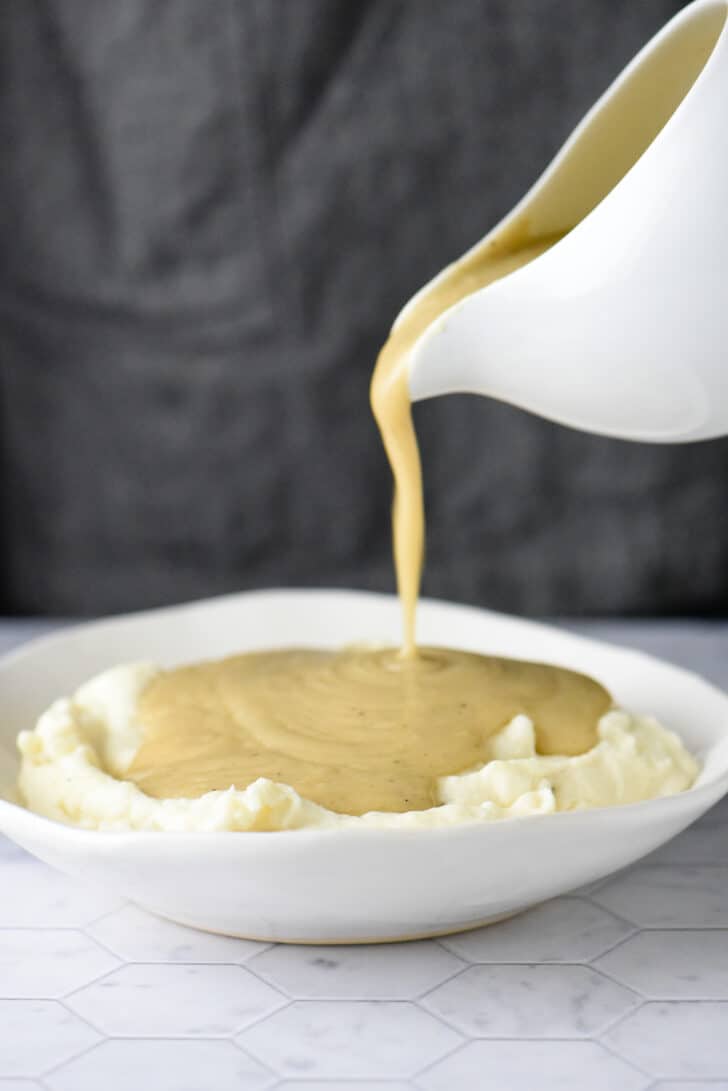 White gravy boat pouring homemade chicken gravy all over a bowl of mashed potatoes.