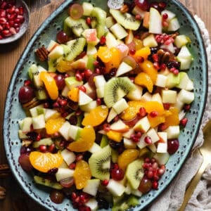 Oval turquoise ceramic bowl filled with Thanksgiving fruit salad made with oranges, grapes, apples, pears, kiwi, pomegranate seeds, dried cranberries and pecans.