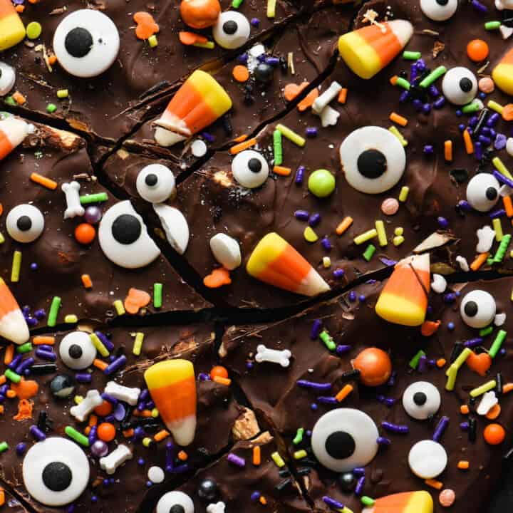 Halloween bark made from chocolate, sprinkles, candy corn and candy eyeballs, broken into pieces, on dark surface.