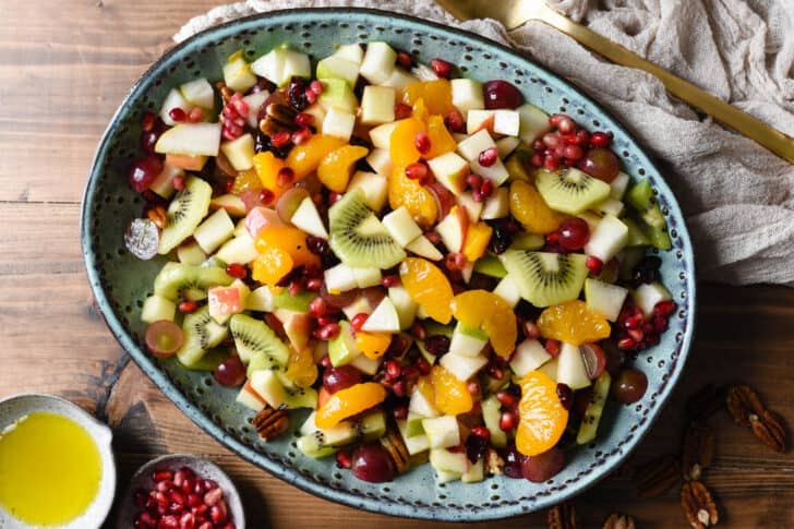 Oval turquoise ceramic bowl with a  mixture of chopped oranges, grapes, apples, pears, kiwi, pomegranate seeds, dried cranberries and pecans.