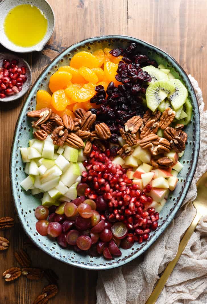Oval turquoise ceramic bowl filled with the ingredients for fall fruit salad, including oranges, grapes, apples, pears, kiwi, pomegranate seeds, dried cranberries and pecans.