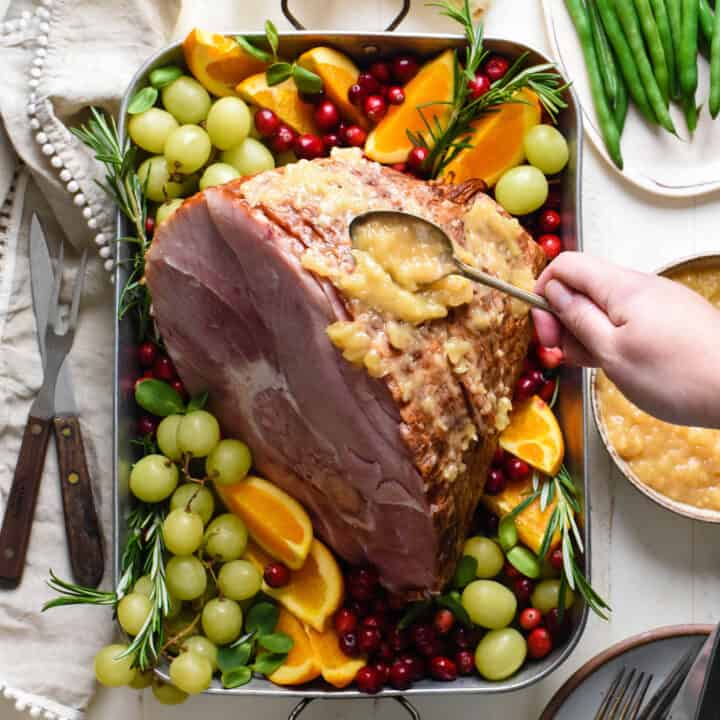 Metal roasting pan with spiral sliced ham surrounded by fruit garnishes, with woman's hand spooning pineapple sauce over ham.