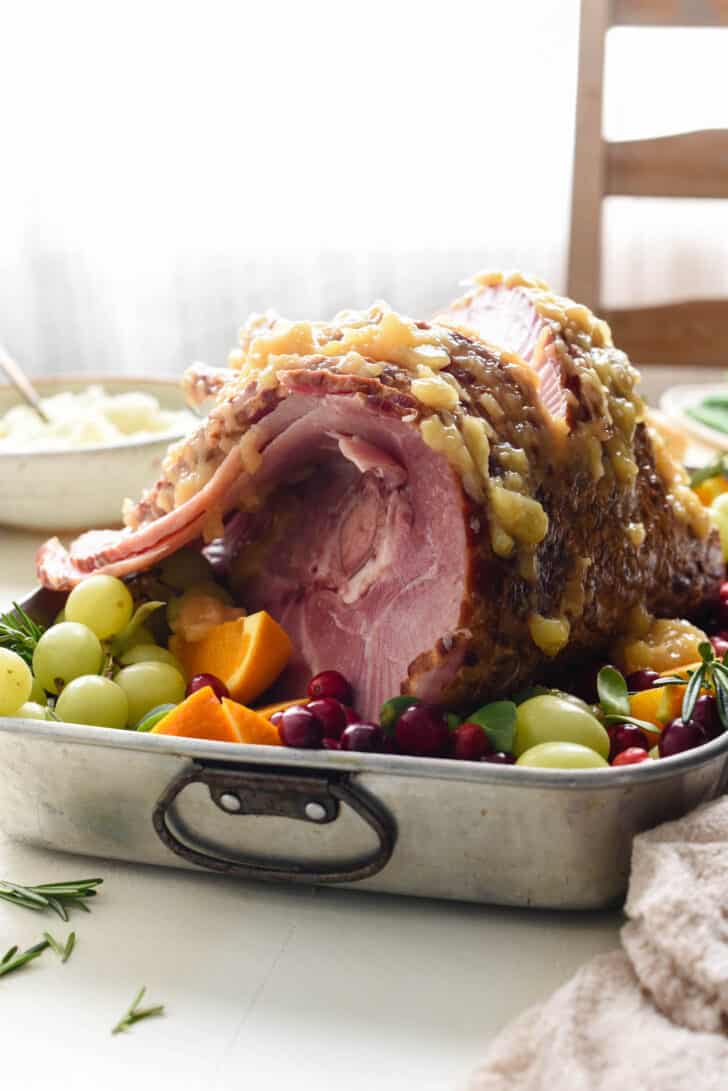 A spiral sliced ham in a roasting pan surrounded by fruit, topped with a pineapple ham glaze.