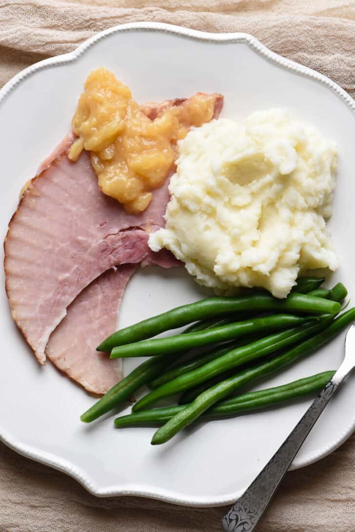 A white plate with mashed potatoes, green beans, and baked ham with pineapple sauce.