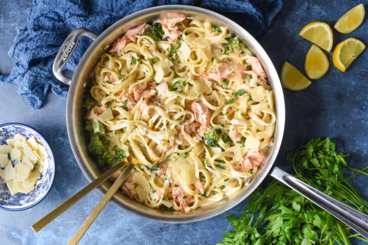 A large stainless steel skillet filled with salmon fettuccine alfredo, garnished with Parmesan cheese, lemon wedges, and parsley.