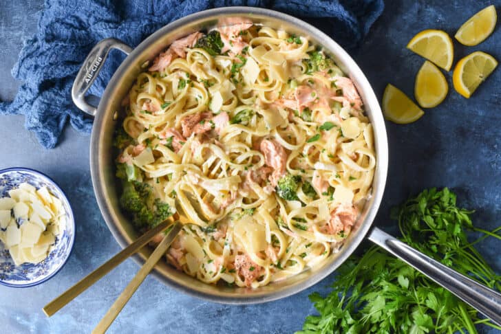 A large stainless steel skillet filled with pasta and fish, with Parmesan cheese, lemon wedges and parsley garnishing the scene.