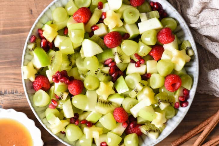 Green grapes, raspberries, pomegranate seeds, kiwi, green apple and star-shaped pineapple pieces tossed together in a gray ceramic bowl.