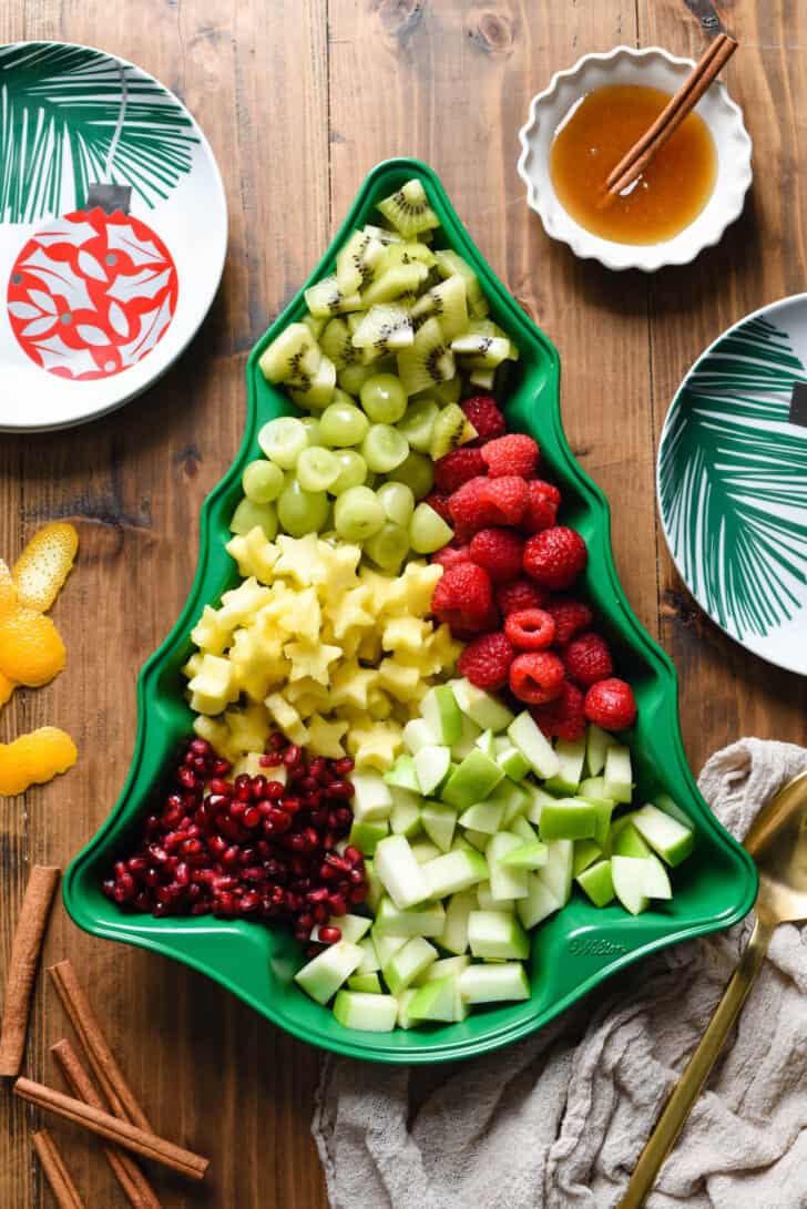 The fixing for holiday fruit salad, including grapes, raspberries, pomegranate seeds, kiwi, pineapple and green apple, arranged in a green Christmas tree shaped baking pan.