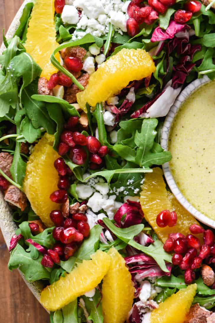 Christmas Salad made with greens, oranges, cheese, pomegranate seeds and nuts, with a bowl of orange salad dressing.