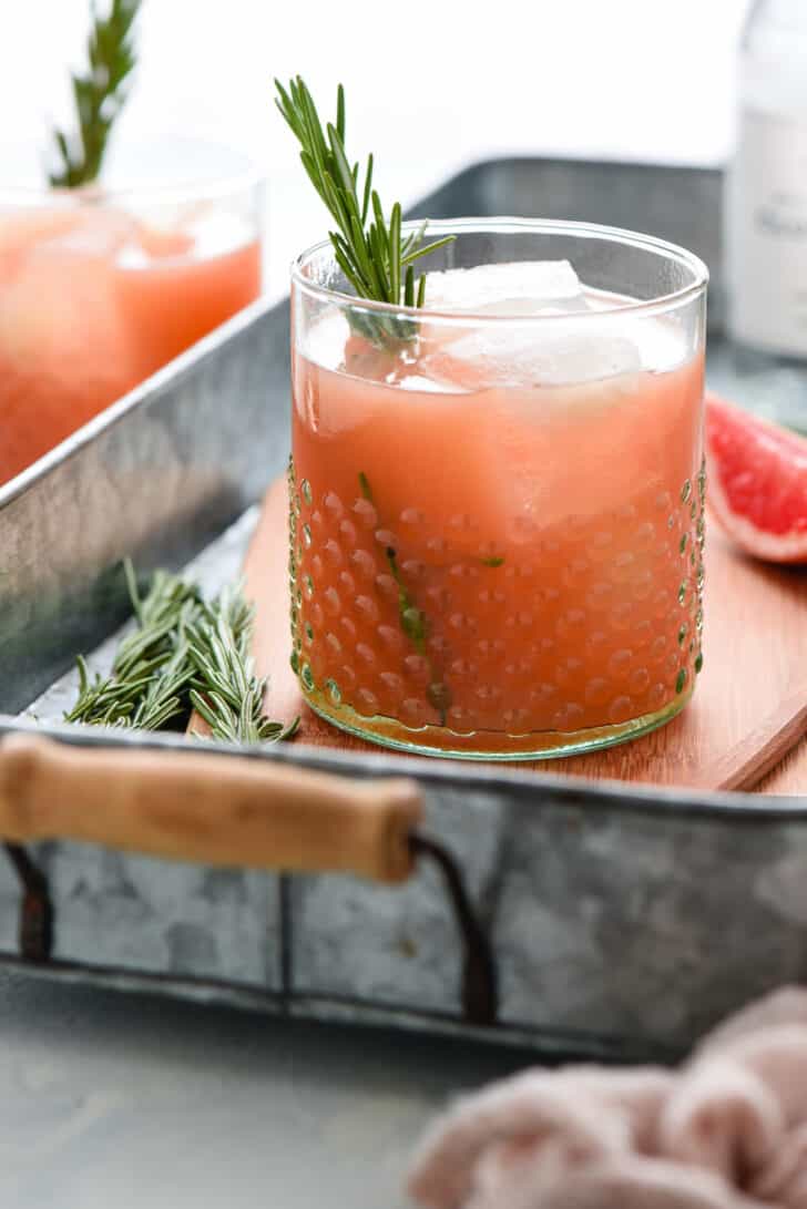 Grapefruit rosemary cocktail with a rosemary sprig garnish in a textured glass on a small wooden cutting board in an aluminum tray.