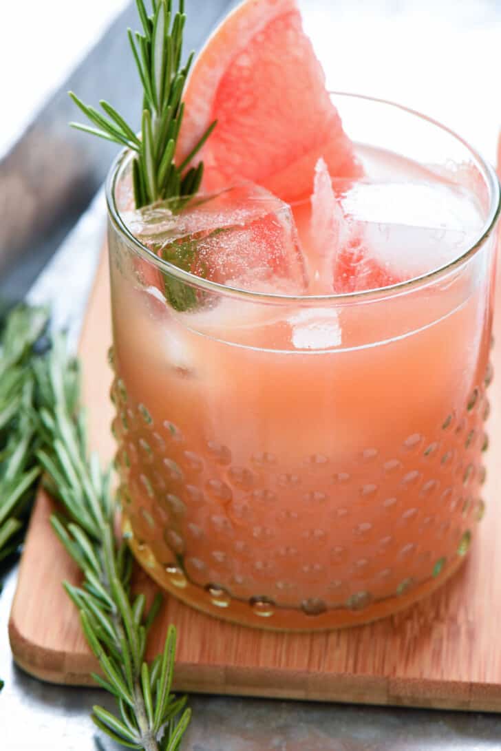 An orange hued drink garnished with a grapefruit wedge and rosemary sprig in a textured glass.