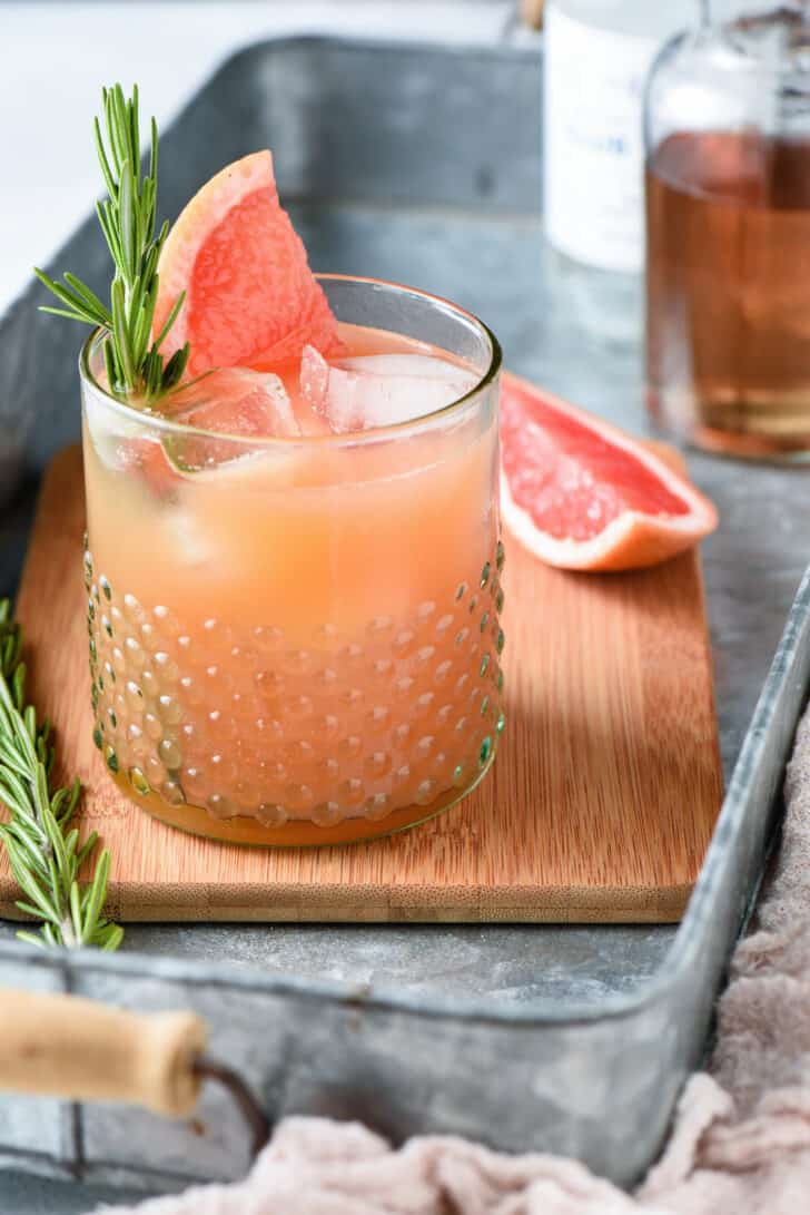 An orange hued drink with a rosemary sprig garnish in a textured glass on a small wooden cutting board in an aluminum tray.