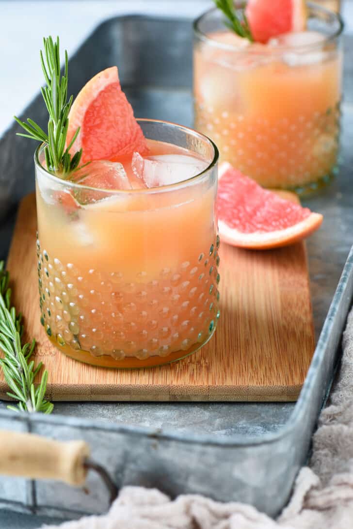 An orange hued grapefruit rosemary cocktail garnished with a grapefruit wedge and rosemary sprig in a textured glass.