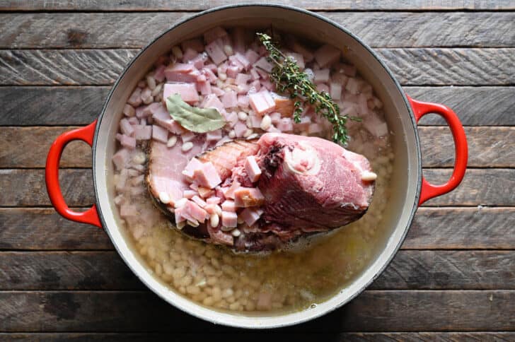 A red Dutch oven filled with mirepoix, water, white beans, herbs and a ham bone.