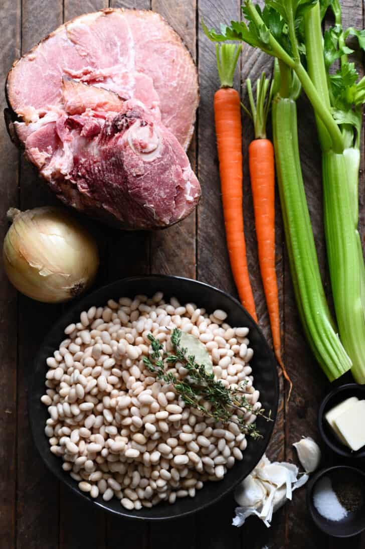Ingredients on a wooden tabletop, including a ham bone, carrots, celery, white beans, butter, garlic, onion and spices.